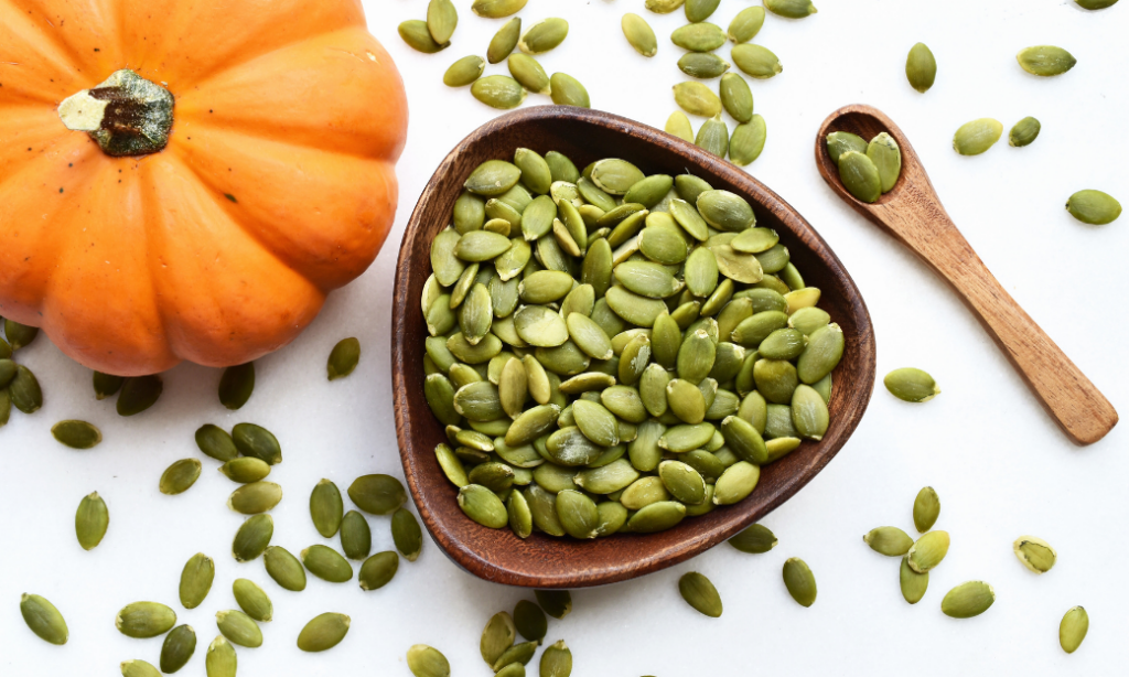 A wooden bowl filled with green pumpkin seeds sits beside a small wooden spoon and an orange pumpkin, with loose seeds scattered on a white surface.