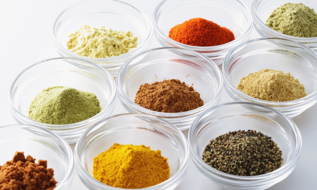 A variety of colorful ground spices displayed in small, clear glass bowls, arranged in a neat grid pattern on a white surface.