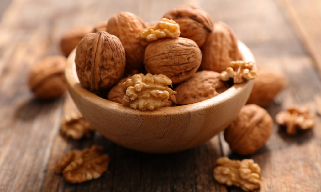 A wooden bowl filled with whole walnuts and walnut halves sits on a rustic wooden surface, with additional walnuts scattered around the bowl.