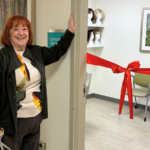 A smiling woman stands by an open door labeled "Consult Room," holding scissors. Inside, a red ribbon is tied across the room, with chairs and wigs visible. Framed artwork hangs on the walls.