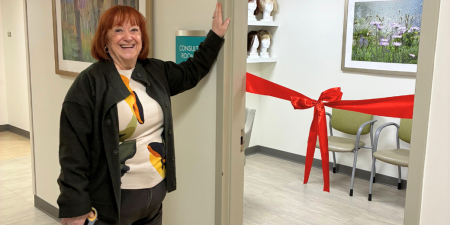 A smiling woman stands by an open door labeled "Consult Room," holding scissors. Inside, a red ribbon is tied across the room, with chairs and wigs visible. Framed artwork hangs on the walls.