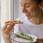 A woman smiling while holding a plate with crispbread topped with cream cheese, cherry tomatoes, and microgreens. She is about to eat one piece, standing by a window with soft natural light.