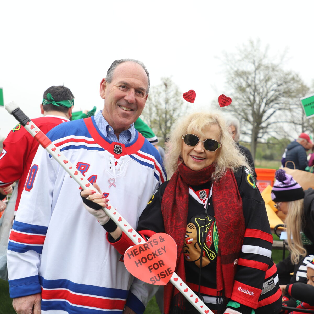 A man and woman smiling outdoors at a hockey-themed event. The man wears a white Rangers jersey and the woman wears a Blackhawks jersey and heart headband. They hold a hockey stick with a heart-shaped sign reading “Hearts & Hockey for Susie.”.