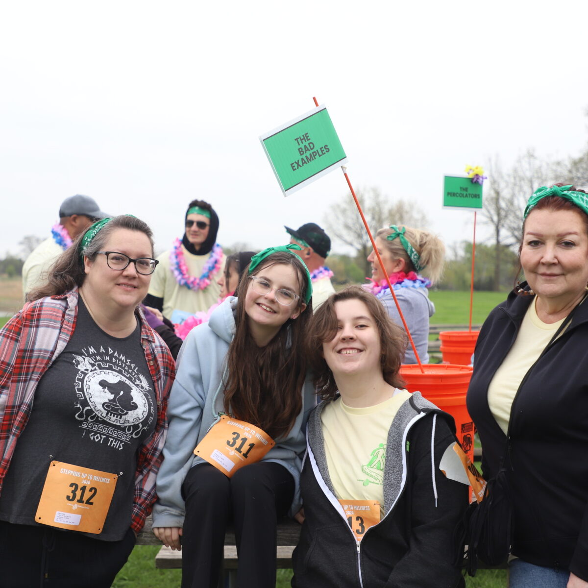 Four people pose and smile outdoors near picnic tables, wearing green bandanas and event bibs. Others sit behind them, some wearing leis. A sign reading “This is fabulous” is held up. The setting is grassy with trees and a lake.