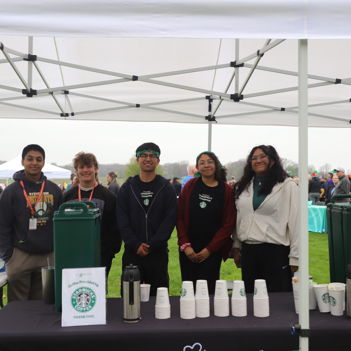 Volunteers stand behind a table offering coffee and water under a white tent. A sign advertises "Coffee Provided by Starbucks Community Champion.