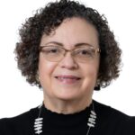 A woman with short curly hair and glasses, wearing a black top and a chunky beaded necklace, smiles at the camera against a white background.