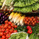 A colorful display of fresh vegetables at a market, including tomatoes, carrots, eggplants, radishes, cilantro, avocados, green beans, and leafy greens neatly arranged in piles.