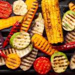 An assortment of colorful vegetables—corn, tomatoes, peppers, squash, eggplant, and zucchini—being grilled, showing char marks and vibrant colors on a barbecue grill.