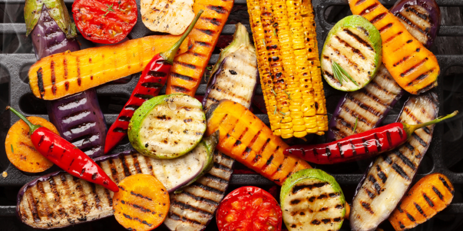 An assortment of colorful vegetables—corn, tomatoes, peppers, squash, eggplant, and zucchini—being grilled, showing char marks and vibrant colors on a barbecue grill.