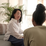 Two women are sitting on a couch in a bright room with a large plant in the background, engaged in a friendly and attentive conversation. One woman faces the camera, smiling, while the other has her back to the viewer.