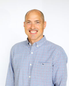 A smiling man wearing a blue and white checkered button-down shirt stands against a plain white background.