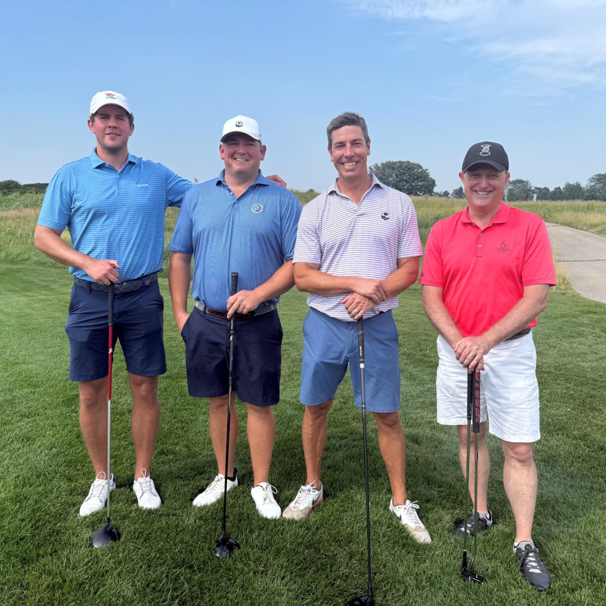 Four men stand on a golf course with their arms around each other, holding golf clubs. They wear polo shirts, shorts, and hats. The sky is clear and grass and a path are visible in the background.