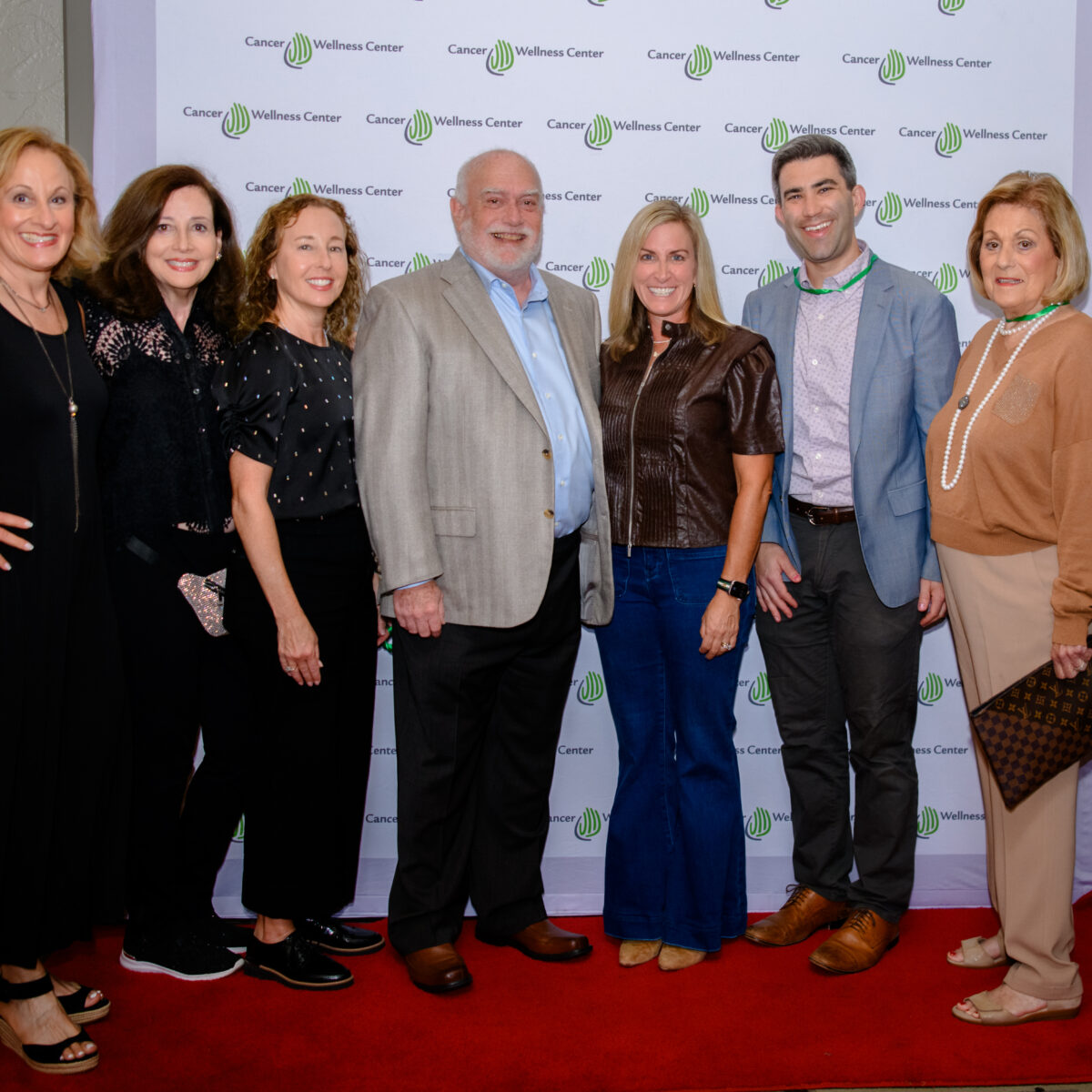Eight adults stand posing and smiling on a red carpet in front of a Cancer Wellness Center step-and-repeat banner. They are dressed in semi-formal attire, with some holding hands or standing close together.