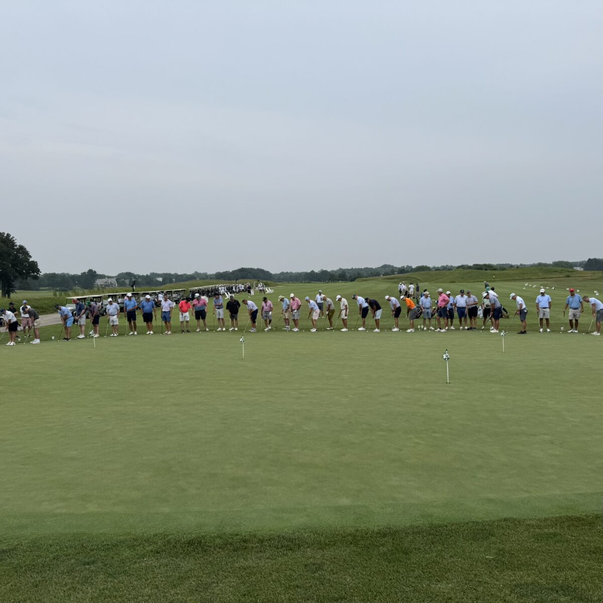 A large group of people stand in a line on a golf course green, bending forward as if preparing to putt, with a cloudy sky and trees in the background.