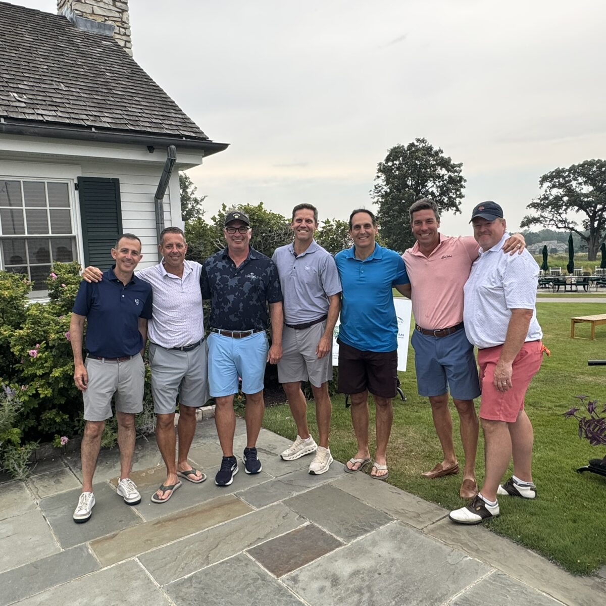 Seven men stand together outdoors on a stone patio, smiling at the camera near a building with green shutters. The background shows grass, trees, and patio furniture under a cloudy sky.
