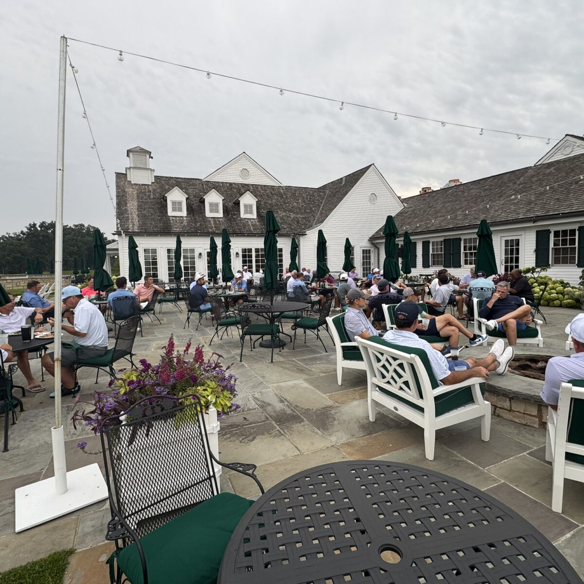 People sit on benches and chairs on a stone patio outside a large clubhouse with white siding and green umbrellas. The scene is relaxed, with many groups socializing under a cloudy sky.
