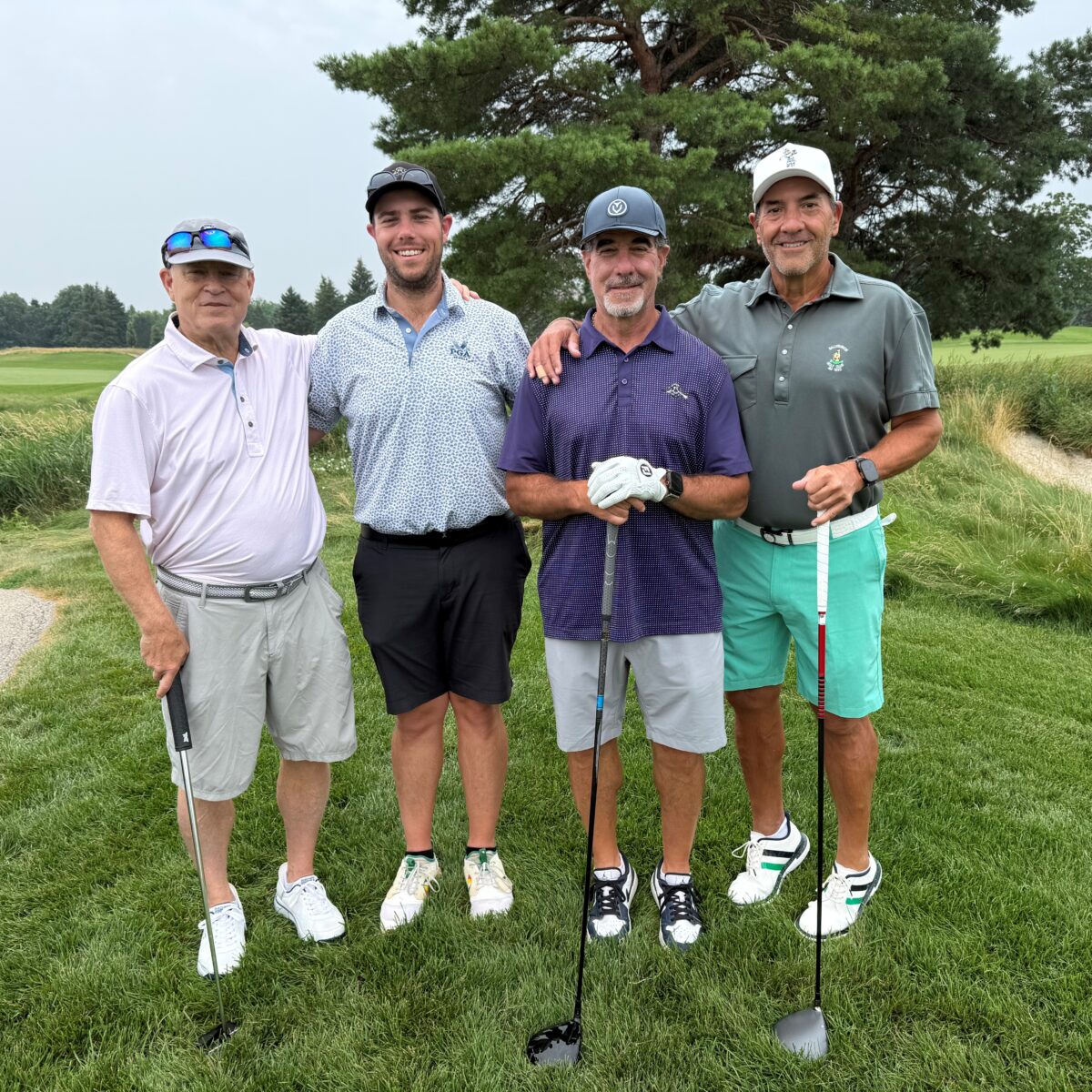 Four men stand on a golf course, each holding a golf club and smiling at the camera. They wear golf attire and hats, with grass and trees visible in the background.