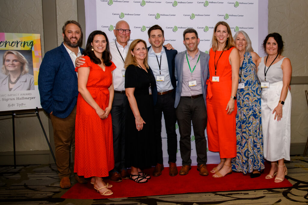 Nine people stand smiling in front of a Wellness Center banner at an indoor event; a sign with a woman's photo and "Honoring" is visible on the left. Everyone is wearing event badges.