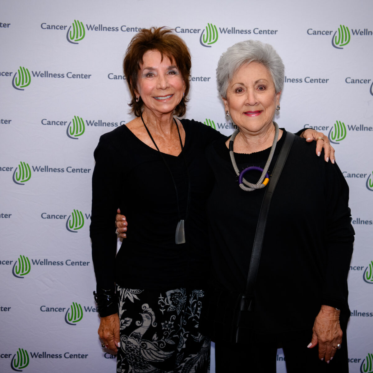 Two older women stand together smiling in front of a Cancer Wellness Center step-and-repeat banner, both dressed in black, one with patterned pants and the other with a statement necklace.