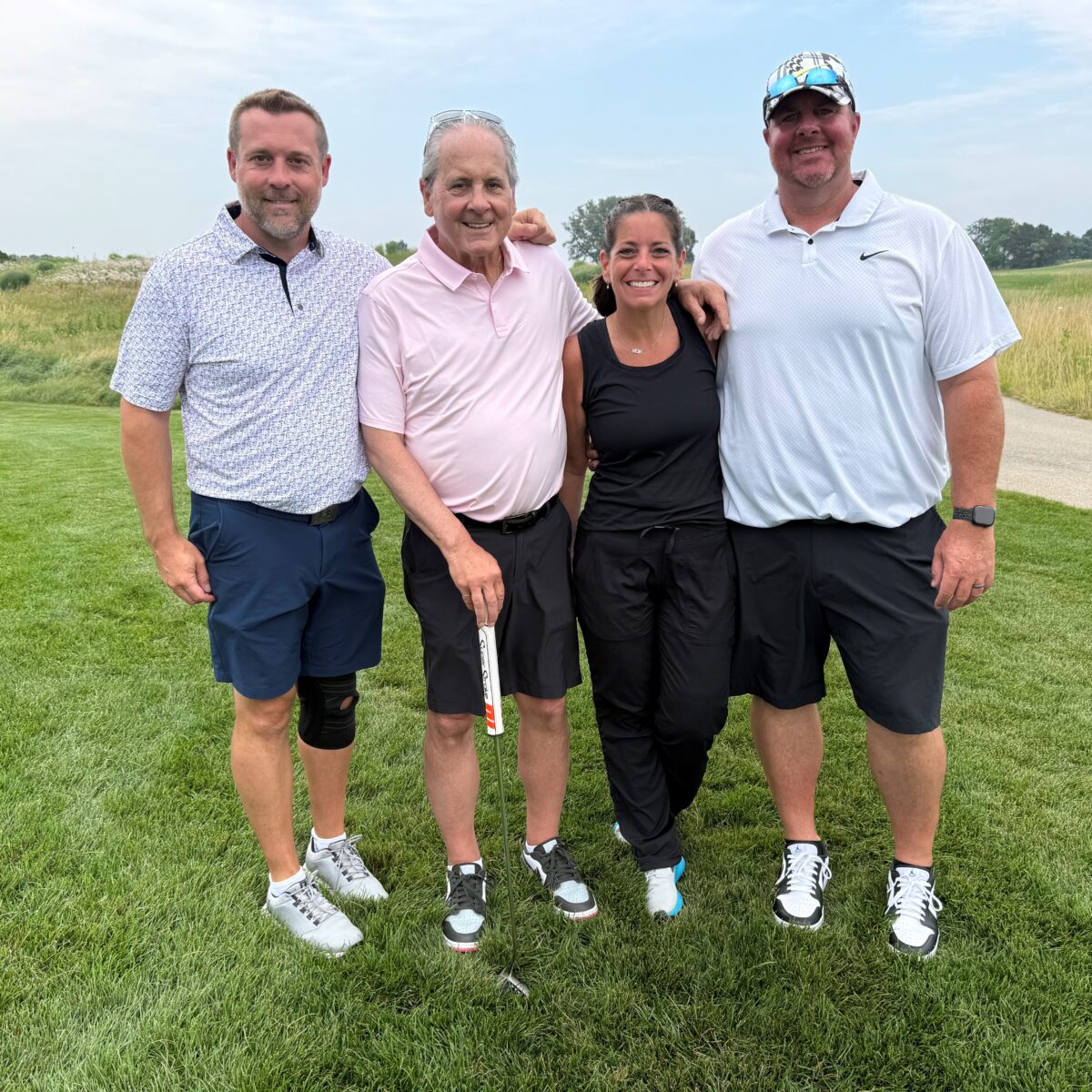 Four adults stand close together on a golf course, smiling at the camera. Three men wear polo shirts and shorts, while the woman wears a sleeveless black top and black pants. Grass and a path are visible in the background.