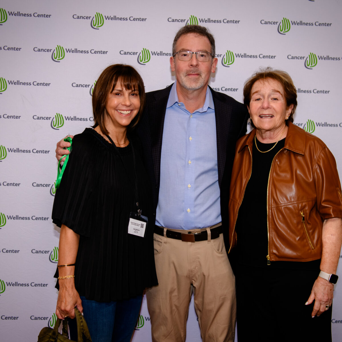 Three adults stand smiling together in front of a Cancer Wellness Center step-and-repeat banner at what appears to be an event. One man is in the center, with two women on either side of him.