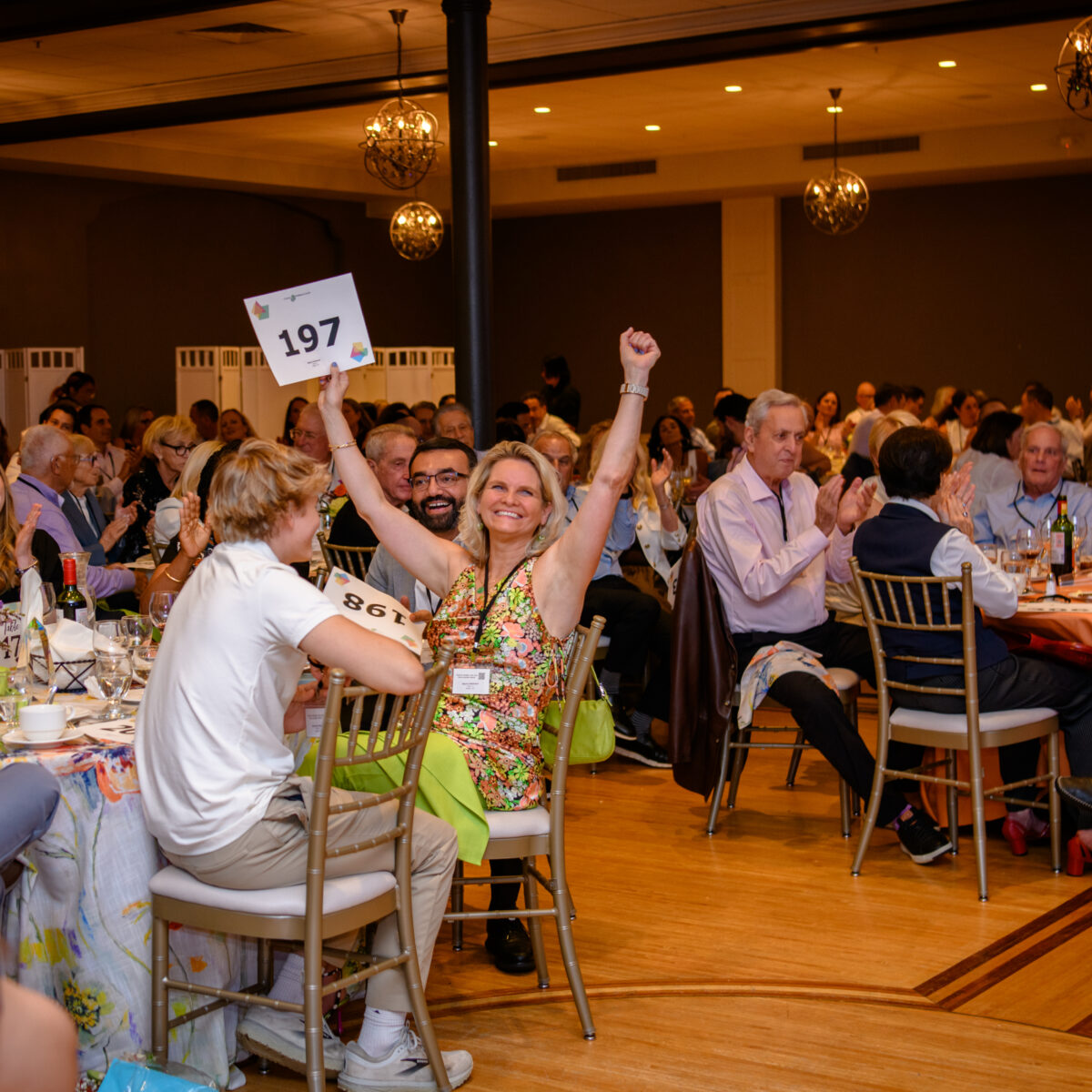 A woman smiles and raises her arms in excitement, holding a “197” sign at an indoor event with round tables and a seated audience clapping.