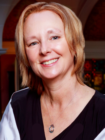 A smiling woman with short, light brown hair wears a black top and a silver necklace, standing indoors with arched architecture and colorful decor in the background.