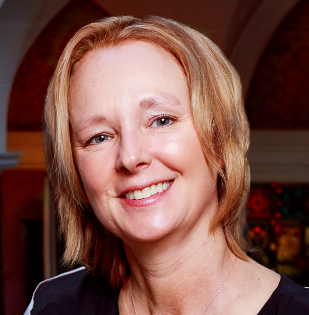 A smiling woman with short, light brown hair wears a black top and a silver necklace, standing indoors with arched architecture and colorful decor in the background.