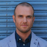 A man with a shaved head and trimmed beard, wearing a light gray blazer over a blue checkered shirt, stands in front of a blue metal roll-up door, looking directly at the camera with a neutral expression.