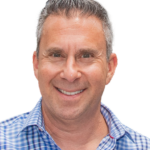 Smiling middle-aged man with short gray hair, wearing a blue and white plaid shirt, standing against a plain white background.