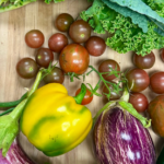 A variety of fresh vegetables on a wooden surface, including green onions, curly kale, cherry tomatoes, yellow bell pepper, striped eggplants, and a jalapeño pepper.