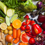 A colorful variety of fresh fruits and vegetables, including oranges, lemons, carrots, broccoli, bell peppers, avocado, blueberries, grapes, potatoes, onions, and leafy greens, arranged on a white surface.