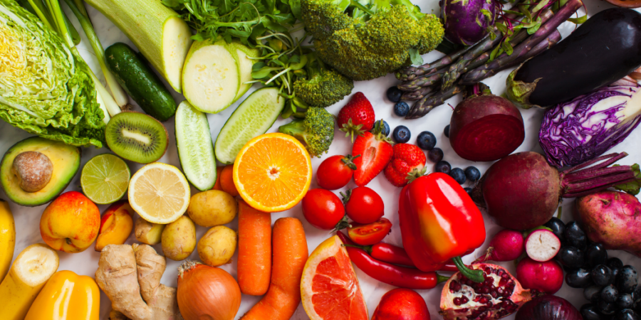 A colorful variety of fresh fruits and vegetables, including oranges, lemons, carrots, broccoli, bell peppers, avocado, blueberries, grapes, potatoes, onions, and leafy greens, arranged on a white surface.