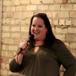 A woman with long dark hair smiles while holding a microphone, standing in front of a brick wall. She is wearing hoop earrings and glasses on her head.